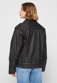 Czarna skórzana kurtka z widocznym szwem, wyprofilowanym kołnierzem i bocznymi kieszeniami na zamek; noszona na jasnoniebieskich dżinsach.