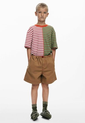 Višebojna prugasta majica s ružičastim i zelenim dijelovima, u kombinaciji s smeđim kratkim hlačama sa crvenim šavovima. Nošeno sa zelenim sandalama i čarapama.