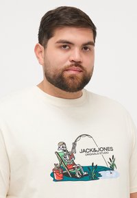 Cremefarbenes T-Shirt mit kurzen Ärmeln, das eine Grafik eines Skeletts zeigt, das aus einem Stuhl angelt, mit dem Text "JACK&JONES ORIGINALS STUDIO".