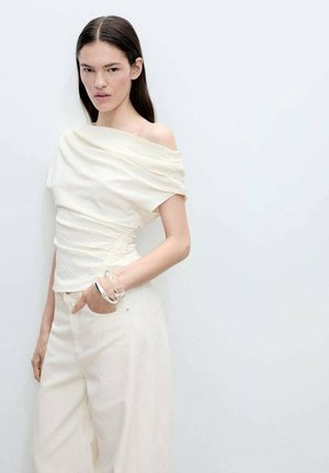 Witte off-the-shoulder top met eenGathered textuur, gecombineerd met lichtgekleurde wijde broek. Model draagt een doorzichtige armband. Eenvoudige achtergrond.
