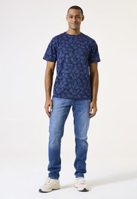 Una camiseta de manga corta de color azul marino presenta un patrón de hojas y un escote redondo. Combinada con jeans de color azul claro y zapatillas beige.