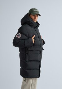 Čierna zateplená bunda s prehnanou kapucňou, na rukáve s logom a so zapínaním na prednej strane s vreckami. Nosená cez svetlé nohavice.