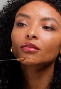 Femme aux cheveux bouclés appliquant un fond de teint liquide sur son menton et son visage à l'aide d'un pinceau de maquillage.