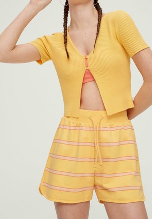 Femme portant un cardigan côtelé jaune à manches courtes sur un crop top orange et un short jaune avec des rayures roses et un cordon, posant avec un bras levé.