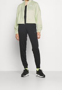 Grön ljusjacka med dragkedja framtill, svarta leggings och svarta sportskor. Jackan har elastiska muddar och en logotyp på bröstet.