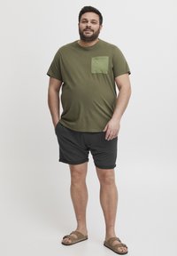Oliven grønn bomulls t-skjorte med lomme foran, kombinert med svarte shorts. Shortsene har rullet kant, og han har på seg brune sandaler.