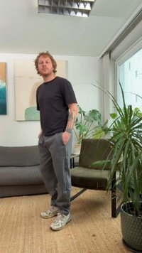 Svart t-skjorte, grå joggebukser og lyse sneakers. Står i et rom med en grønn plante, grå sofa og abstrakt veggkunst.