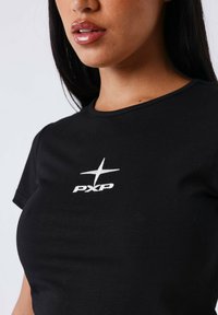 Vrouw in een zwart shirt met een witte ster en het "PXP"-logo op de borst, met glanzende lippen en lang donker haar.