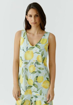 Femme aux cheveux bruns mi-longs portant une robe sans manches bleu clair avec un motif de citrons jaunes et de feuilles vertes, debout devant un fond uni.