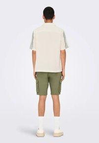 Camisa de manga corta en color crema con paneles laterales estampados, combinada con pantalones cortos cargo en color oliva que cuentan con múltiples bolsillos. Calcetines blancos texturizados y zapatos.