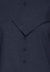 Cardigan bleu marine avec une texture côtelée, fermeture boutonnée à l'avant et col plié. La manche est étendue, mettant en valeur l'élasticité du tissu.