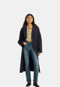 Gabardina marina con un diseño de doble botonadura, top corto beige claro, jeans azules y botas negras hasta el tobillo. Tela suave y corte estructurado.