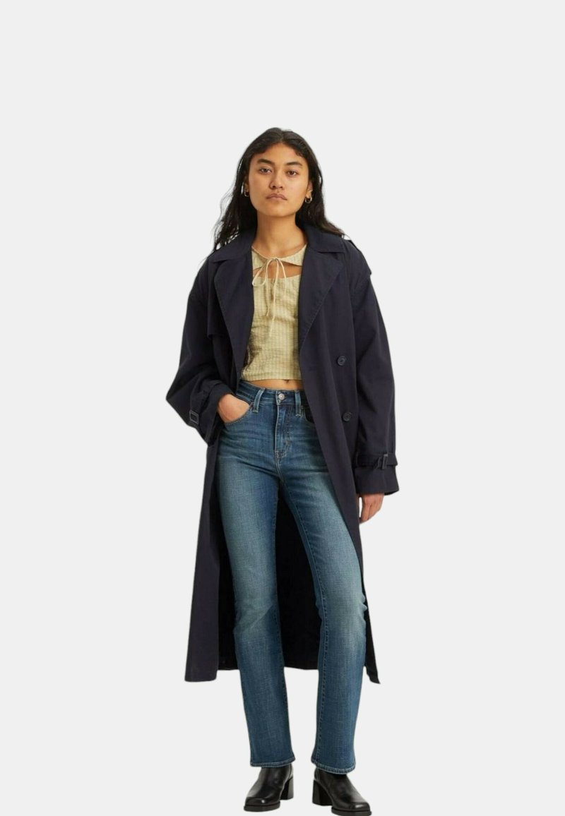 Gabardina marina con un diseño de doble botonadura, top corto beige claro, jeans azules y botas negras hasta el tobillo. Tela suave y corte estructurado.