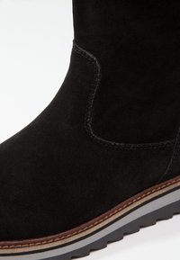Bottine en daim noir avec coutures apparentes, bordure marron, et semelle à crampons superposée en gris et noir.