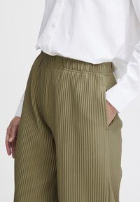 Pantaloni plissettati verde oliva con cintura elastica, tasche e una texture liscia, abbinati a una camicia bianca con bottoni.