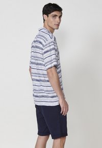 Pánska košeľa s krátkymi rukávmi s horizontálnymi námorníckymi a bielymi pruhmi, vyrobená z ľahkého materiálu. V kombinácii s námorníckymi kraťasmi s rolovaným lemom.