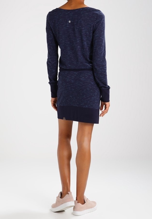 Personne debout, dos à la caméra, portant une robe bleu marine à manches longues et des baskets rose clair sur un fond blanc uni.