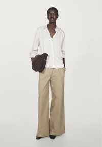 White button-up shirt with long sleeves, paired with wide beige trousers. Model holds a dark brown suede bag, featuring a soft texture.