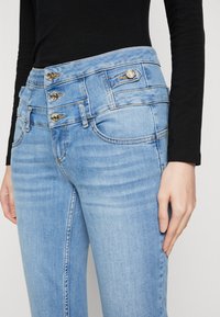 Gai zili džinsi ar augstu jostasvietu, trīs pogu aizdari un kabatām, ar smalkām nodiluma pazīmēm un oranžiem šuvēm.