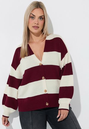 Kvinde med lige blondt hår iført en vinrød og hvid stribet knappet cardigan og mørke jeans, der står foran en hvid baggrund.
