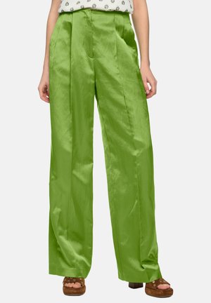 Frau trägt hoch taillierte, weit geschnittene Satin-Hosen in Limettengrün mit Falten, kombiniert mit braunen offenen Plateau-Sandalen und einer gemusterten, in die Hose gesteckten Bluse.