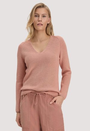 Frau mit blondem Haar, die einen pinkfarbenen, gestrickten Pullover mit V-Ausschnitt und pinke Hose mit Kordelzug trägt, steht mit einer Hand in der Tasche vor einem schlichten Hintergrund.