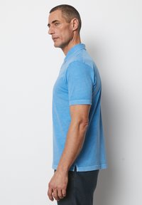 Helles blaues Poloshirt mit kurzen Ärmeln und Kragen, das eine weiche Textur und ein leicht meliertes Muster aufweist. Seitenschlitze am Saum.