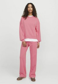 Roza kapuca in usklajene hlače iz mehkega materiala, ki se ponašajo z udobnim krojem in dolgimi rokavi. Pod njimi je vidna bela majica.