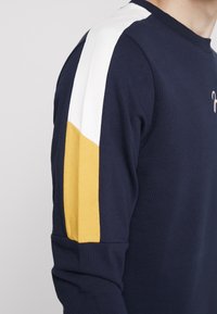Felpa blu con maniche a blocchi di colore bianco e giallo, colletto a coste e texture morbida. Presenta un piccolo logo sul petto.