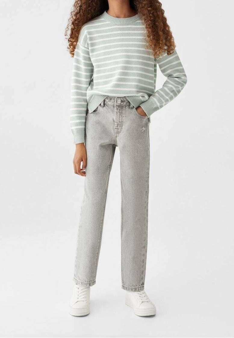 Person klädd i en ljusgrön och vit randig tröja, ljusgrå högmidjade jeans, vita sneakers, med ena handen i fickan.