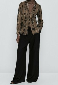 Ruskea nappikauluspaita, jossa on musta abstrakti kuvio, yhdistettynä väljiin mustiin housuihin, korostaen rentoa istuvuutta ja kevyttä kangasta.