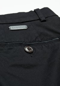 Zbliżenie na kieszeń czarnych spodni z guzikem i metalową metką wygrawerowaną napisem "bugatti" na pasku.