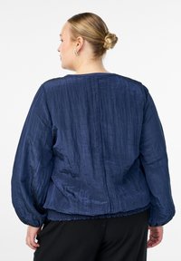 Ženska z blond lasmi v speti pričeski, oblečena v guban temno modri dolg rokav in črne hlače, obrnjena stran od bele ozadja.