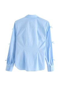 Camicia di cotone azzurro con maniche lunghe, polsini con bottoni e dettagli annodati sulle maniche. Presenta un colletto e un orlo curvo sul retro.