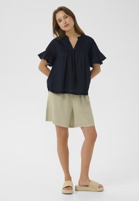 Blusa blu marina con maniche corte a volant e scollatura a V, abbinata a pantaloni corti beige chiaro con tasche frontali e vestibilità rilassata.