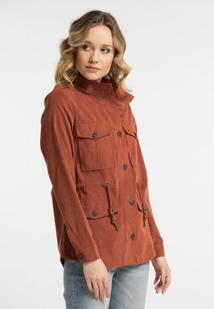 Donna con capelli biondi mossi che indossa una giacca color ruggine con quattro tasche frontali e jeans azzurri, che guarda di lato.