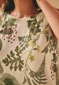 Femme portant une robe sans manches à imprimé botanique vert et jaune, tenant ses cheveux, avec un bracelet en or au poignet.