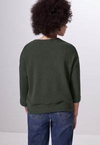 Mörkgrön, ribbad stickad tröja med bred halsringning, trekvartsärmar och en avslappnad passform, i kombination med blå jeans.