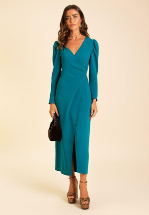 Teal wikkeljurk met lange mouwen, diepe V-hals, geplooide schouders en een split aan de voorkant. Gecombineerd met bruine plateauhakken en een zwarte clutch.