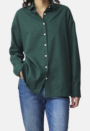 Camicia verde con bottoni realizzata in tessuto morbido, dotata di un colletto classico, maniche lunghe e bottoni bianchi; indossata sopra jeans blu.