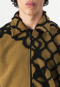 Brown fleece jacket with a textured black geometric pattern, featuring a zippered front and a soft collar. Adjustable fit and uncluttered design.