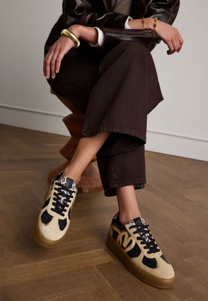 Baskets beiges et noires avec une surface texturée, arborant un logo sur le côté, des lacets noirs et un col à motif. Design à semelle plate.