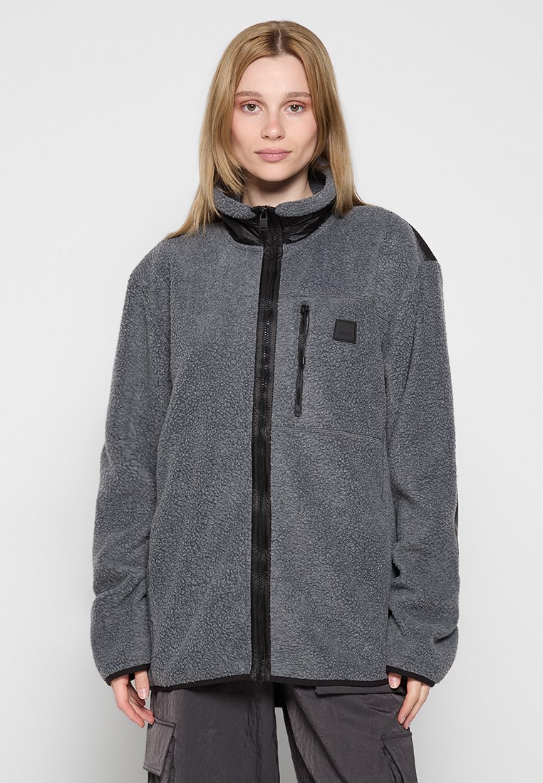 Veste en polaire gris foncé avec un col haut, fermeture éclair à l'avant et une poche sur la poitrine gauche. Présente des accents noirs sur les épaules et l'ourlet.