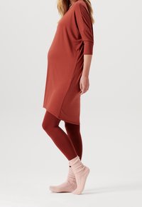 Rostfarbendes, locker sitzendes Umstandskleid mit 3/4-Ärmeln, kombiniert mit kastanienbraunen Leggings und rosafarbenen gerippten Socken. Glatte Stoffoberfläche.