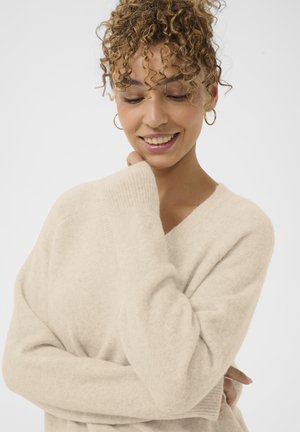 Femme aux cheveux bouclés portant des boucles d'oreilles créoles en or et un pull beige, souriant doucement les yeux fermés sur un fond blanc uni.
