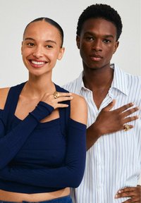 Deux personnes posent ensemble. L'une porte un haut côtelé bleu marine à épaules dénudées, l'autre une chemise blanche à rayures. Toutes deux arborent des bagues dorées distinctives.