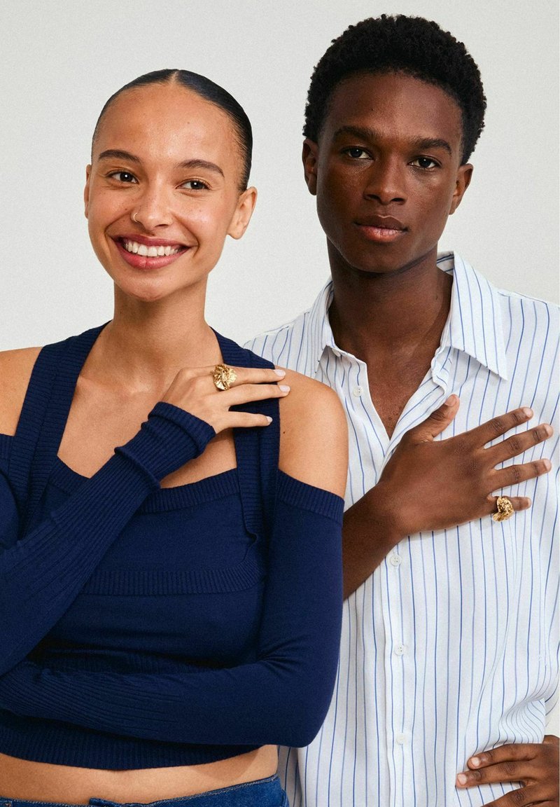 Deux personnes posent ensemble. L'une porte un haut côtelé bleu marine à épaules dénudées, l'autre une chemise blanche à rayures. Toutes deux arborent des bagues dorées distinctives.