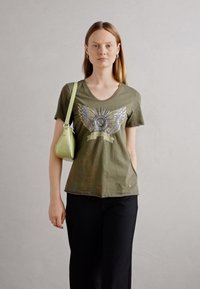 Olijfgroene V-hals t-shirt met metallic vleugelgrafiek, gecombineerd met een gestructureerde lichtgroene handtas. Zwarte broek maakt de outfit compleet.