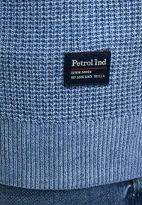 Pulover tricotat de culoare albastră deschisă, cu un model texturat și tiv ondulat. Etichetă neagră cu textul "Petrol Ind" și "DENIM DRIVEN" imprimat pe ea.