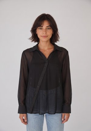 Joven mujer con cabello oscuro y corto lleva una camisa negra transparente con botones y vaqueros azul claro, de pie frente a un fondo liso de color gris claro.